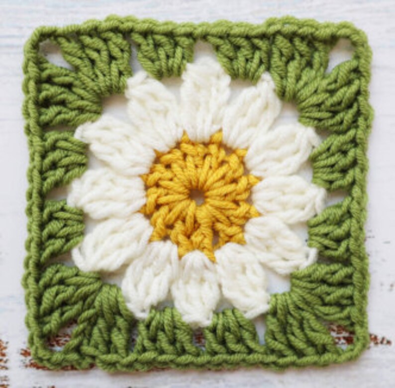 May include: A crocheted square featuring a white daisy flower in the centre. The flower has a yellow centre and green leaves around the edges.