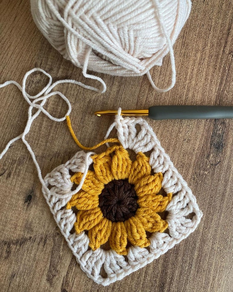 May include: A crocheted sunflower square with a brown centre and yellow petals. The square is surrounded by a white crocheted border. A crochet hook and yarn are also visible in the image.