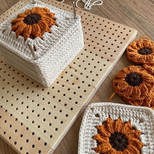 May include: A wooden pegboard with holes for holding yarn and crochet projects. There are three crocheted sunflower squares on the board, two are finished and one is in progress.