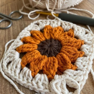 May include: A crocheted sunflower with orange petals and a brown centre. The sunflower is surrounded by a white crocheted border.