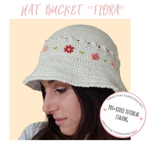 Può includere: Cappello bianco a secchiello in crochet con fiori ricamati in rosa e rosso. Il cappello è indossato da una persona con lunghi capelli scuri. Il testo "PDF+VIDEO TUTORIAL ITA/ENG" è su un cerchio rosa con un cuore.