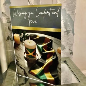 May include: A card with the words "Wishing you Comfort and peace" is displayed next to a lit candle with a Jamaican flag design. A black, yellow, and green ribbon is draped nearby.
