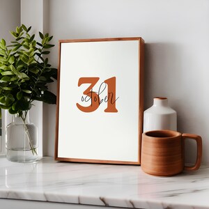 Puede incluir: Una impresión enmarcada con el número 31 en color naranja quemado y la palabra "Octubre" escrita en letra cursiva negra. El marco es de madera marrón claro.