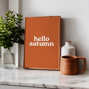 Può includere: Una stampa incorniciata con sfondo arancione e testo bianco che dice "hello autumn".