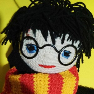 Puede incluir: Muñeca de crochet con cabello negro, gafas redondas y una bufanda a rayas rojas y amarillas. La muñeca lleva un atuendo de crochet negro.