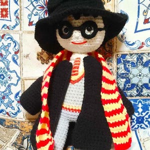 May include: Crocheted doll wearing a black hat, glasses, a black coat, and a red and yellow striped scarf.