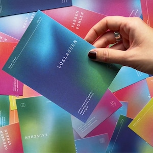 May include: A hand holding a card with the word "LOSLASSEN" in white text, with a gradient of blue, green, and purple. Other cards with similar designs and text are scattered around, featuring various colors and words.