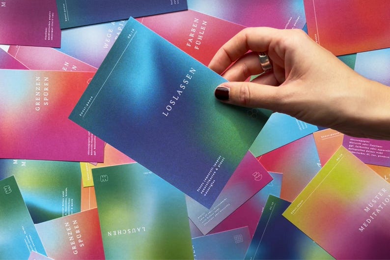 May include: A hand holding a card with the word "LOSLASSEN" in white text, with a gradient of blue, green, and purple. Other cards with similar designs and text are scattered around, featuring various colors and words.