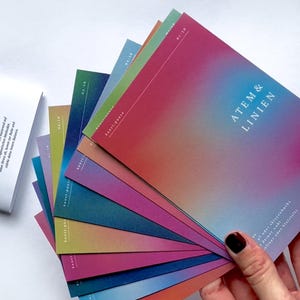 May include: A collection of colorful cards with a gradient design, fanned out to display their vibrant hues. The cards feature the text "ATEM & LINIEN" and "kunst:pause" on a white background. The cards are held by a person.