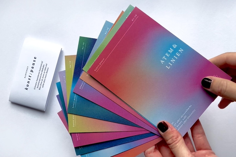 May include: A collection of colorful cards with a gradient design, fanned out to display their vibrant hues. The cards feature the text "ATEM & LINIEN" and "kunst:pause" on a white background. The cards are held by a person.