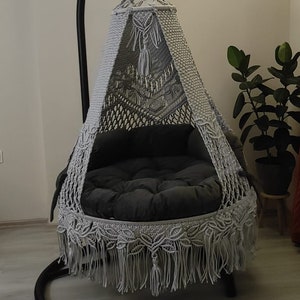 May include: A grey macrame hanging chair with a black metal stand. The chair has a black cushion and a fringe trim.