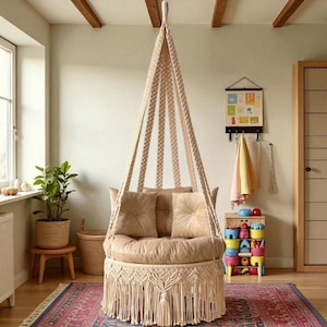 Puede incluir: Una silla colgante de macramé beige con asiento redondo y flecos en la parte inferior. La silla tiene dos cojines cuadrados en el respaldo y está suspendida del techo. La habitación tiene una alfombra, una planta y una puerta de madera.