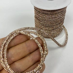 May include: A spool of rose gold rhinestone trim. The trim is made of small, round rhinestones that are sewn together to create a continuous chain. The trim is wound around a white spool.