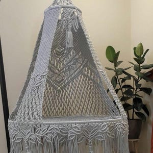 Könnte beinhalten: Ein weißer Makramee-Hängesessel mit Fransenbesatz und Quasten. Der Sessel hängt an einem schwarzen Metallständer.