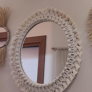 Könnte beinhalten: Ein runder Spiegel mit einem weißen Makramee-Rahmen. Der Rahmen hat einen Fransenrand und ein gewebtes Muster.