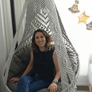 May include: A grey macrame hanging chair featuring an intricate knotted pattern, a conical top with a decorative bow, and a circular base. The chair includes a plush, dark grey circular cushion designed to fit within the woven frame structure.