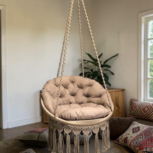 Op de afbeelding: Een beige macramé hangstoel met een gecapitonneerd kussen. De stoel hangt aan het plafond met dikke, geweven touwen. De stoel heeft een ronde zitting en franjes aan de onderkant. Decoratieve kussens liggen op de vloer.