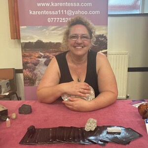 May include: A woman wearing glasses and a black vest top sits at a table with a stack of tarot cards, crystals, and a sign that reads "KAREN-TESSA SPIRITUAL MEDIUM www.karen-tessa.com karen-tessa111@yahoo.com 0777252"