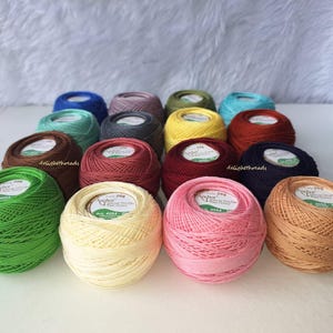May include: A collection of colorful Anchor brand crochet thread balls in various shades, including green, yellow, pink, and blue. Each ball is labeled with the brand name and product information. The threads are neatly arranged, showcasing a variety of crafting colors.
