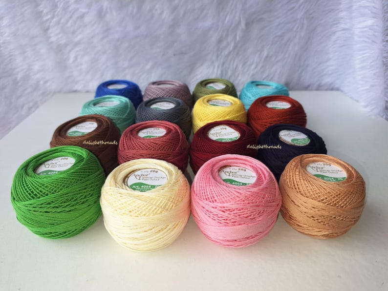 May include: A collection of colorful Anchor brand crochet thread balls in various shades, including green, yellow, pink, and blue. Each ball is labeled with the brand name and product information. The threads are neatly arranged, showcasing a variety of crafting colors.
