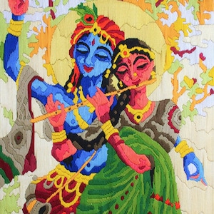 Op de afbeelding: Een kleurrijk geborduurd kunstwerk dat een stel, mogelijk Krishna en Radha, in een liefdevolle omhelzing toont. De man speelt fluit terwijl de vrouw een bloem vasthoudt. Het kunstwerk is geplaatst tegen een achtergrond van bomen en bloemen.