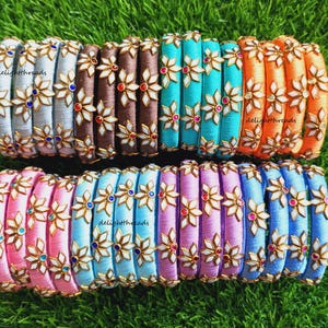 Puede incluir: Una colección de coloridas pulseras de hilo de seda. Las pulseras están adornadas con diseños florales y piedras preciosas. Los colores incluyen rosa, azul, morado, marrón, naranja y turquesa. Las pulseras se exhiben sobre hierba verde.
