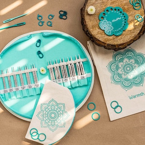 Peut inclure: Un ensemble d'aiguilles à tricoter de couleur turquoise avec différentes tailles, des marqueurs de mailles et un gabarit. L'ensemble comprend un étui rond, une petite pochette et un sac en toile, tous ornés d'un motif floral. Des accessoires turquoise supplémentaires sont dispersés.