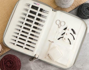 Puede incluir: Un juego de agujas de tejer intercambiables KnitPro en un estuche de tela con cremallera. El set incluye varios pares de puntas de metal negro, cables circulares, un par de tijeras pequeñas y una cinta métrica cuadrada blanca.