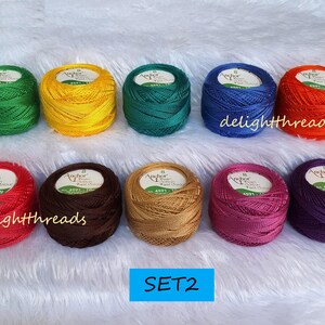 May include: A collection of ten spools of Anchor Pearl Cotton thread in various colors, including green, yellow, teal, blue, orange, red, brown, gold, pink, and purple. The spools are arranged on a white, fluffy surface.