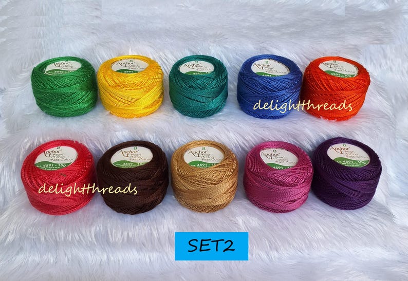 May include: A collection of ten spools of Anchor Pearl Cotton thread in various colors, including green, yellow, teal, blue, orange, red, brown, gold, pink, and purple. The spools are arranged on a white, fluffy surface.