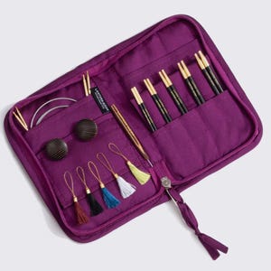 May include: A purple zippered knitting needle case, open to reveal various knitting tools. The case contains several black and gold knitting needles, stitch markers, and other accessories. The case is made of a quilted fabric and has a matching zipper pull.