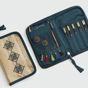 May include: A teal fabric case with a zipper closure, featuring a tan front panel with three embroidered dark floral motifs. The interior contains slots for knitting needles, two wooden buttons, and five small colorful hanging tassels.