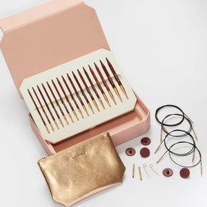 May include: A knitting needle set in a pink box, with a gold pouch. The set includes multiple wooden needles with gold tips, black cables, and various accessories. The pouch has the text "Knit Pro" in gold.