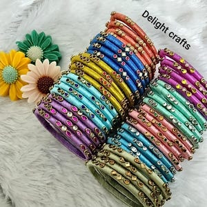 Op de afbeelding: Een verzameling kleurrijke armbanden in roze, blauw, groen, geel en paars. Elke armband is versierd met decoratieve versieringen en geplaatst tegen een witte, zachte achtergrond. De tekst "Delight crafts" is zichtbaar.