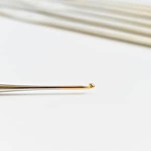 Peut inclure: Gros plan sur un crochet doré avec un manche argenté. La pointe du crochet est incurvée et pointue, conçue pour tirer le fil à travers les boucles. Plusieurs autres crochets sont légèrement visibles en arrière-plan.
