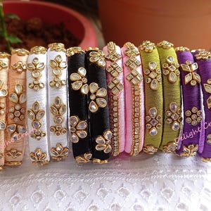 Puede incluir: Una colección de pulseras coloridas, cada una envuelta en hilo de seda y adornada con adornos dorados y piedras preciosas transparentes. Las pulseras son en tonos melocotón, blanco, negro, rosa, verde y morado, expuestas sobre una superficie blanca.