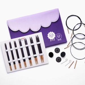 May include: A knitting needle set with black and gold needles, black cables, and accessories. The set includes a purple case with a scalloped edge and sheep graphics. The needles are displayed on a white holder.