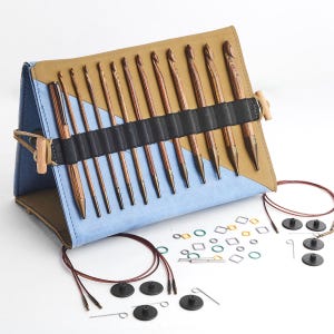 Puede incluir: Un juego de ganchillos con ganchos de madera y una funda azul y beige. El juego incluye ganchos de varios tamaños, marcadores de punto y otros accesorios. La funda se asegura con un cierre de palanca de madera.