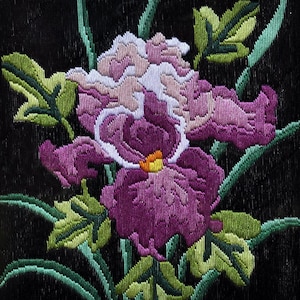 Könnte beinhalten: Detaillierte Stickerei einer Irisblüte in Lila, Weiß und Gelb auf schwarzem Hintergrund. Grüne Blätter und Stiele umgeben die Blüte und erzeugen ein lebendiges, strukturiertes Blumenmuster.