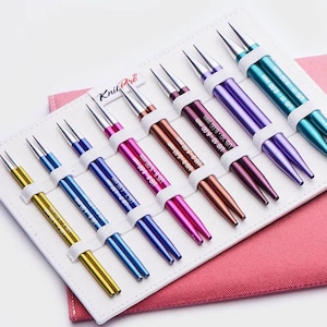 May include: A collection of Knit Pro circular knitting needles in a range of colours, including yellow, blue, pink, and purple. The needles are stored in a white case with a pink fabric backing. Each needle is labelled with its size in US and mm.
