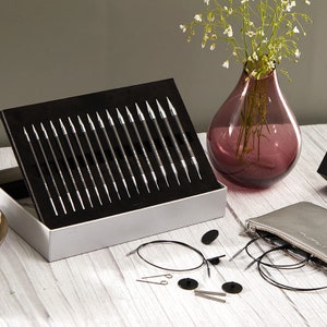 Peut inclure: Un ensemble d'aiguilles à tricoter dans une boîte noire et argentée. Les aiguilles ont des pointes argentées et des manches noirs. Des accessoires de tricot, tels que des câbles et des marqueurs de mailles, sont disposés autour de la boîte. Un vase violet avec des fleurs blanches est en arrière-plan.