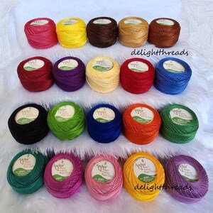 Puede incluir: Una colecci&oacute;n de ovillos de hilo Anchor Pearl Cotton de colores vibrantes, incluyendo rosa, amarillo, marr&oacute;n y azul. Cada ovillo est&aacute; etiquetado con una etiqueta del producto. Los hilos est&aacute;n dispuestos sobre una superficie blanca y esponjosa.
