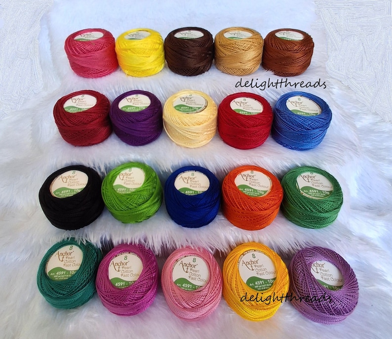 Puede incluir: Una colecci&oacute;n de ovillos de hilo Anchor Pearl Cotton de colores vibrantes, incluyendo rosa, amarillo, marr&oacute;n y azul. Cada ovillo est&aacute; etiquetado con una etiqueta del producto. Los hilos est&aacute;n dispuestos sobre una superficie blanca y esponjosa.