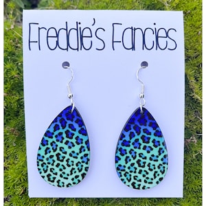 May include: A pair of teardrop earrings with a blue and teal leopard print design. The earrings are suspended from silver-colored hooks. The earrings are displayed on a white card with the text "Freddie's Fancies".