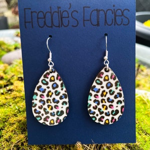 May include: Teardrop-shaped earrings with a colorful leopard print pattern. The earrings are white with black, blue, yellow, and pink spots. The earrings hang from silver hooks and are displayed on a navy blue card with the text "Freddie's Fancies".