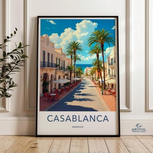 May include: A framed art print of Casablanca, Morocco. The image depicts a sunny street lined with palm trees, cafes, and buildings. The sky is blue with fluffy white clouds. The word "CASABLANCA" is printed at the bottom.