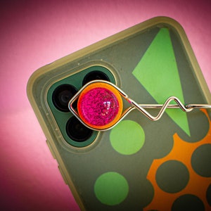 Könnte beinhalten: Eine rosa und orangefarbene Handyhülle mit einem silbernen Handyringhalter. Die Handyhülle hat ein geometrisches Muster mit grünen und orangen Kreisen.