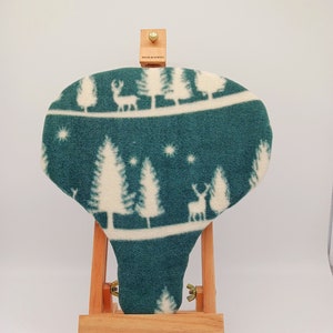 May include: A teal blue fleece bike seat cover with a white winter scene design featuring trees and deer. The cover is displayed on a wooden easel.