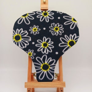 May include: A black bicycle seat cover with a white daisy pattern and yellow centers. The seat cover is displayed on a wooden easel.