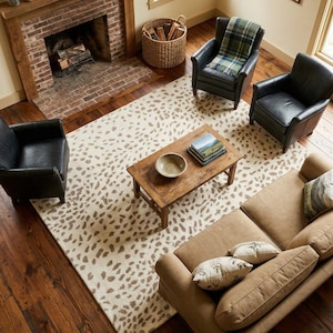 May include: A living room arrangement featuring a cream-colored area rug with a brown speckled pattern, a rectangular wooden coffee table with a bowl and books, three black leather armchairs, and a tan sofa with two patterned throw pillows.